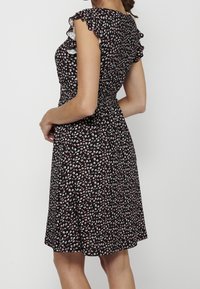 Femme portant une robe noire avec un petit motif floral blanc et rose, des manches courtes à volants et une taille élastiquée, vue de dos.