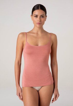 Donna con capelli scuri che indossa un top aderente rosa senza maniche e biancheria intima beige, in piedi davanti a uno sfondo chiaro e uniforme.