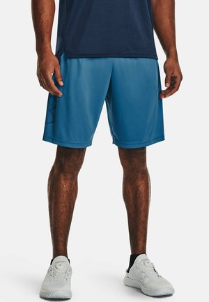 Man met een marineblauw shirt, blauwe sportshorts en witte sneakers, staand recht vooruit tegen een effen lichte achtergrond.