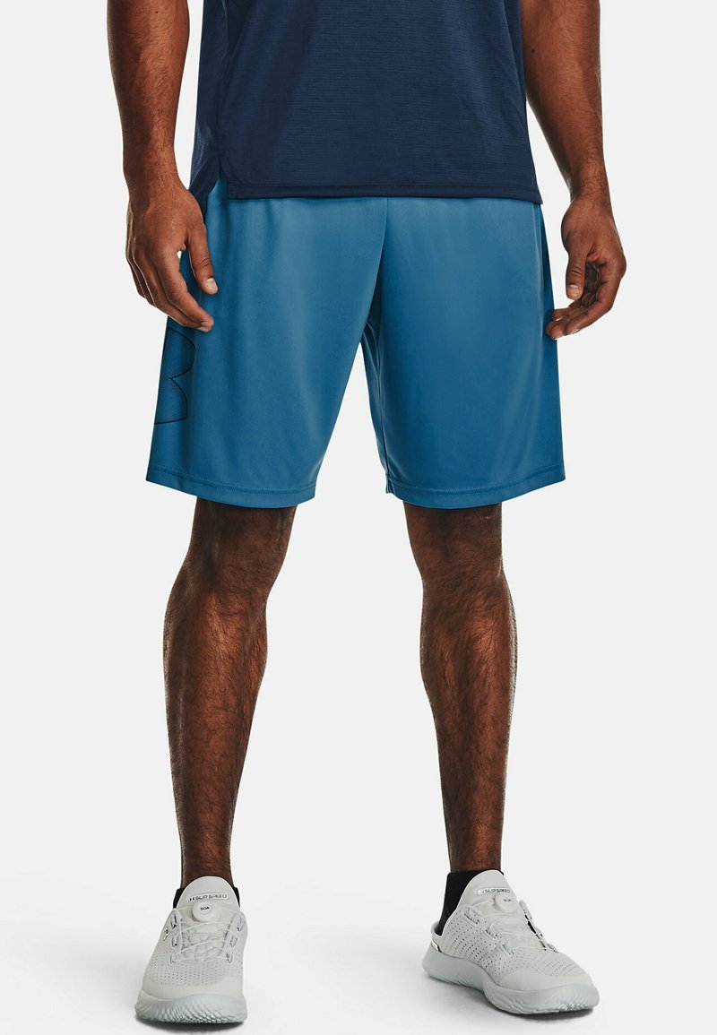 Homme portant une chemise bleu marine, un short de sport bleu et des baskets blanches, debout face à un fond clair et uni.