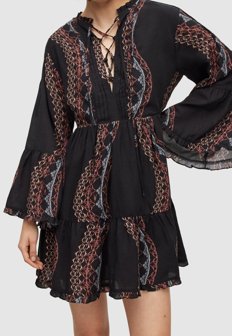 Femme portant une robe noire à manches longues avec des volants aux poignets et à l'ourlet, avec des motifs verticaux rouges et blancs en forme de chaîne et un col en V lacé.