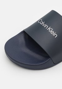 Infradito blu navy realizzate in plastica testurizzata, con una larga fascia con il marchio "Calvin Klein" in rilievo. Suola piatta con finitura liscia.