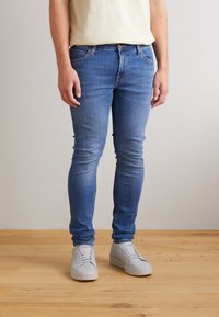 Jeans ajustados de mezclilla en azul claro, con bolsillos delanteros, cierre de cremallera y un sutil efecto desteñido. Combinados con zapatillas grises con cordones.