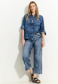 Denim shirt in donkerblauw met knopen aan de voorkant en twee borstzakken, gecombineerd met lichtblauwe acid-wash broek met wijdere pijpen en zwarte sandalen.