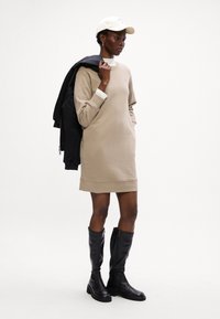 Beige Sweatshirtkleid mit langen Ärmeln und Bündchen, kombiniert mit schwarzen kniehohen Stiefeln und einer weißen Mütze, lässiger Stil mit minimalistischem Design.