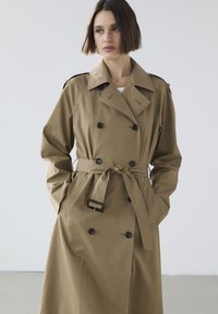 Khakifarbener Trenchcoat mit doppelter Knopfleiste, schwarzen Knöpfen und einem taillierten Gürtel. Hergestellt aus glattem Stoff und mit Schulterklappen ausgestattet.