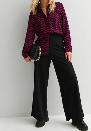 Frau trägt eine magentafarbene Bluse mit schwarzen Herzen, schwarze weite Hosen, schwarze klobige Stiefel und hält eine schwarze Tasche mit goldener Kette.