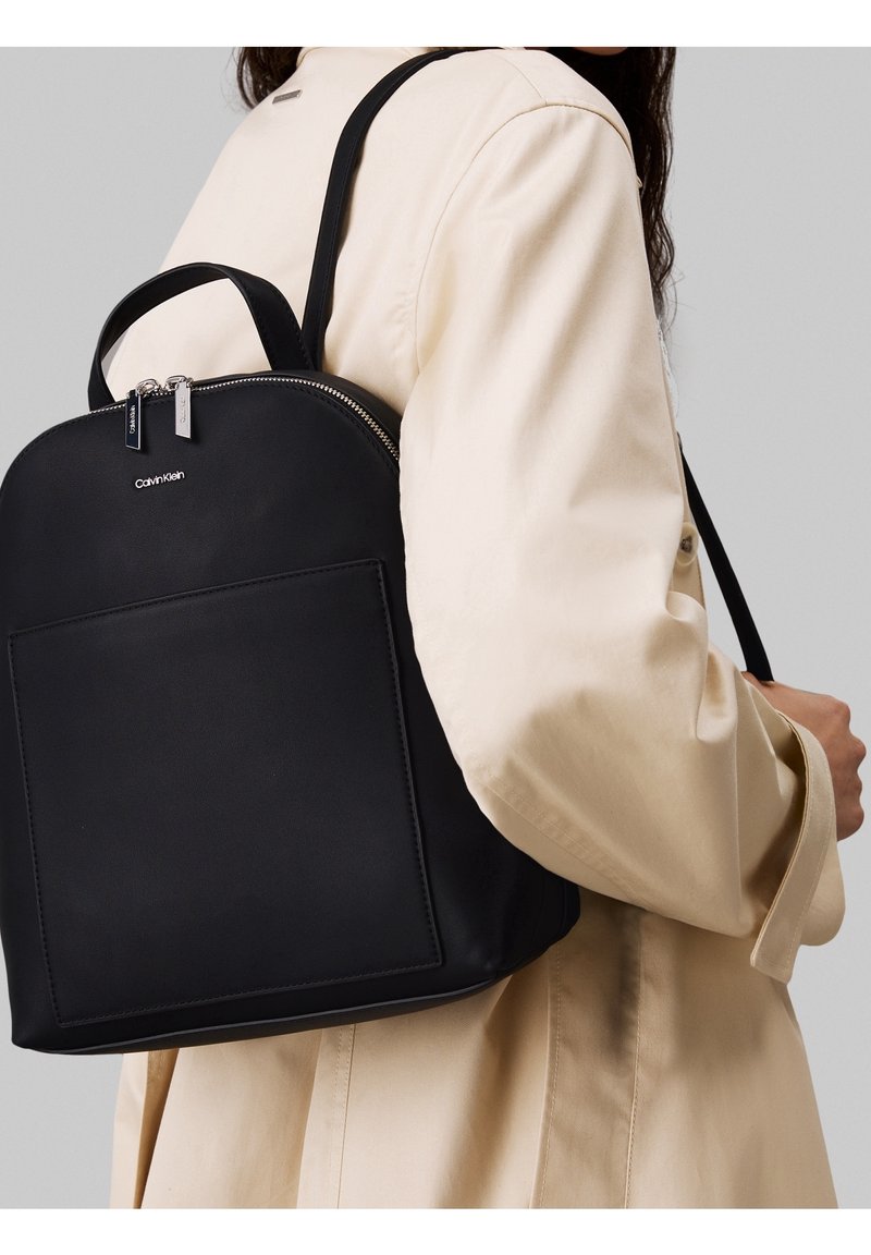 Mochila negra hecha de tela lisa con parte superior redondeada, que cuenta con un bolsillo frontal y detalles de cremallera plateada. Llevada sobre un abrigo color crema.