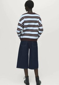 Persona vista da dietro che indossa un maglione a righe blu e marroni, pantaloni cropped larghi scuri e stivaletti neri.