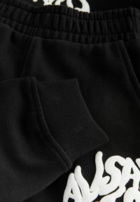 Čierne bavlnené tepláky s rebrovaným manžetovým zakončením a elastickým pásom. Výrazný biely grafický text na látke.