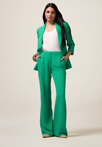 Turquoise blazer en uitlopende broek set gemaakt van gladde stof. Witte top eronder, geaccessoriseerd met een gouden armband.