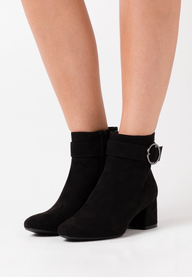 Jambes portant des bottines en daim noir à talons blocs avec des sangles à boucle argentée sur fond blanc.