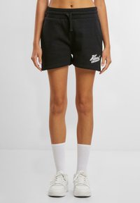 Schwarze Baumwollshorts mit elastischem Taillengummi und Kordelzug, mit weißer Logo-Stickerei am linken Saum. Getragen mit weißen Socken und Turnschuhen.