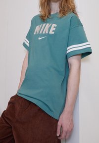 Teal bomull T-shirt med vita axelrandar, med "NIKE" logotyp i grått och en liten Nike swoosh. Bärs med bruna kordfläskbyxor.