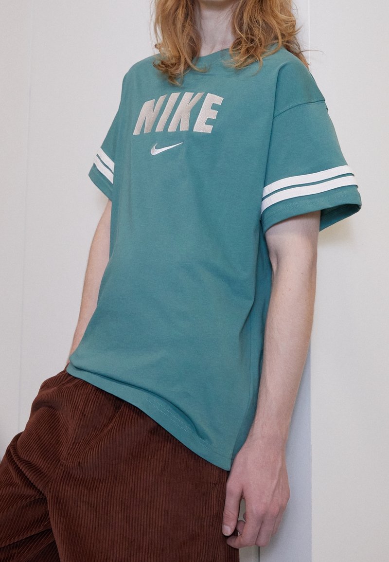 Teal bomull T-shirt med vita axelrandar, med "NIKE" logotyp i grått och en liten Nike swoosh. Bärs med bruna kordfläskbyxor.