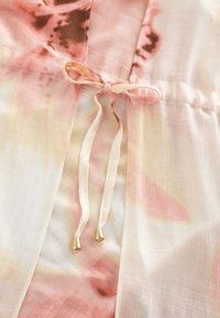 Close-up of a pink and cream tie-dye fabric with a tied bow and two hanging strings with gold metal tips.