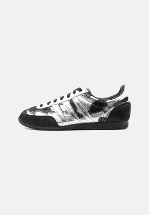 Χαμηλό sneaker με μεταλλικό ασημί επάνω μέρος και μαύρες σουέντ λεπτομέρειες, με μαύρα κορδόνια και υφασμένο πέλμα σε λευκό φόντο.