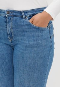 Jean en denim bleu à coupe slim. Les caractéristiques incluent une poche avant, une fermeture à bouton en métal et des coutures visibles le long des seams.