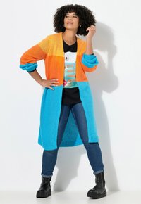 Cardigan lungo colorato con un design a gradiente arancione e blu. Tessuto in maglia, apertura frontale e maniche arrotolate abbinate a una maglietta nera e jeans.