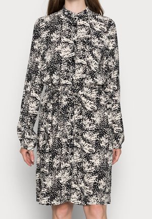 Femme portant une robe à manches longues à motif abstrait noir et blanc, avec une ceinture à la taille et un col boutonné.