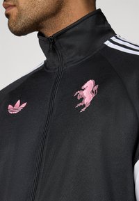 Μαύρο μπουφάν με φερμουάρ, με ροζ κεντητό λογότυπο αλόγου στο στήθος και ροζ λογότυπο Adidas στην αριστερή πλευρά. Διαθέτει λευκές ρίγ ες στους ώμους.