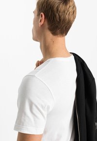 T-shirt bianca in cotone con scollatura rotonda, maniche corte e cuciture visibili; il modello tiene una giacca nera sulla spalla.