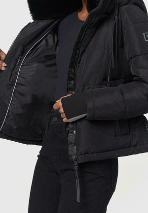 Personne portant une veste matelassée noire avec capuche doublée de fourrure, montrant l'intérieur ouvert et les détails de la fermeture éclair, associée à une chemise et un pantalon noirs.