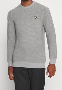 Grå strikket sweater med horisontalt ribbet tekstur, rund halsudskæring og lille gul broderet logo på brystet, kombineret med mørke bukser.