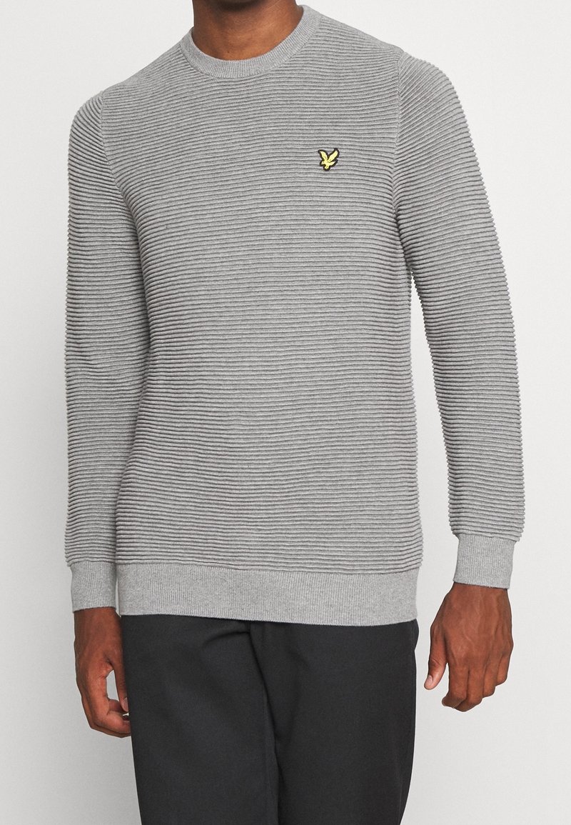 Grå strikket sweater med horisontalt ribbet tekstur, rund halsudskæring og lille gul broderet logo på brystet, kombineret med mørke bukser.