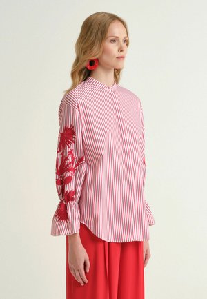 Donna che indossa una camicetta a righe rosse e bianche con ricami floreali rossi sulle maniche, pantaloni rossi a gamba larga e grandi orecchini rossi, rivolta verso destra.