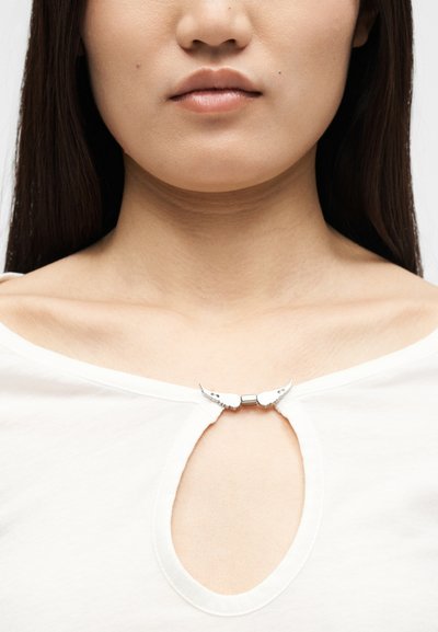 Κοντινό πλάνο ενός ατόμου που φοράει λευκό τοπ με ντεκολτέ σε σχήμα κλειδαρότρυπας, στερεωμένο με ένα ασημένιο διακοσμητικό κούμπωμα σε σχήμα φτερού.