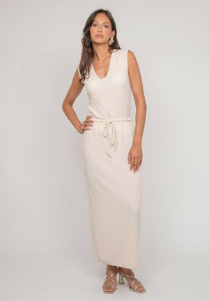 Femme portant une robe maxi crème sans manches avec une ceinture nouée à la taille et des sandales à talons dorées à brides, debout devant un fond uni.
