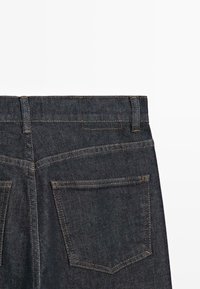 Jeans in denim indaco scuro vista posteriore che mostrano la parte superiore con due tasche posteriori e cuciture arancioni a contrasto su sfondo bianco.