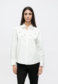 Weißes Langarmhemd mit Knopfleistenkragen, vorne mit silbernen Blumenverzierungen. Kombiniert mit schwarzen Hosen.