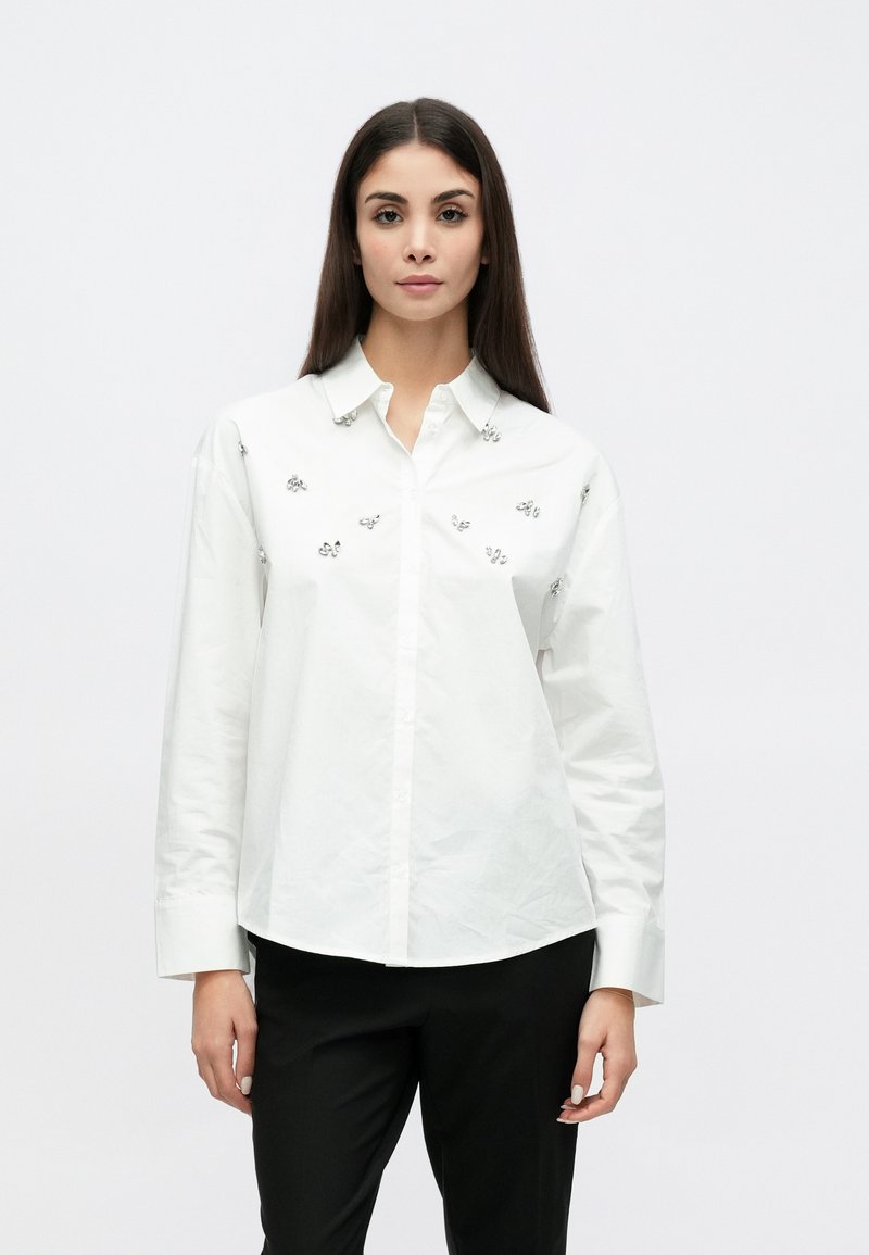 Weißes Langarmhemd mit Knopfleistenkragen, vorne mit silbernen Blumenverzierungen. Kombiniert mit schwarzen Hosen.