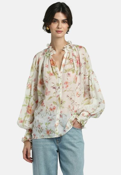 Kvinde iført gennemsigtig hvid bluse med blomsterprint, flæsekant ved kraven og manchetter, kombineret med lyseblå jeans, stående med den ene hånd i lommen.