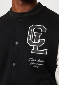 Černá varsity bunda s bílým logem "CL" a textem. Vyrobena z jemné tkaniny s žebrovaným límcem, předními cvoky a kontrastními rukávy.