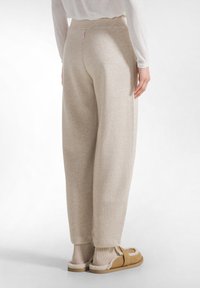 Helles Beige, glitzernde Sweatpants mit weitem Bein und elastischem Bund, von hinten gezeigt. Kombiniert mit beigen Hausschuhen und gerippten Socken.