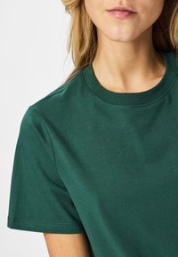 Blond persoon die een effen donkergroen T-shirt met korte mouwen en ronde hals draagt, gecropt van schouders tot lippen tegen een witte achtergrond.
