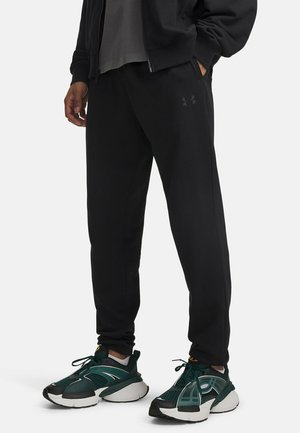 Personne portant un pantalon de jogging noir Under Armour avec les mains dans les poches et des chaussures de sport vertes et blanches, debout sur un fond uni.