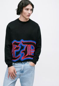 Maglione nero con una grande grafica colorata in rosso e blu. Collo e polsini a costine. Indossato con jeans azzurri chiari.