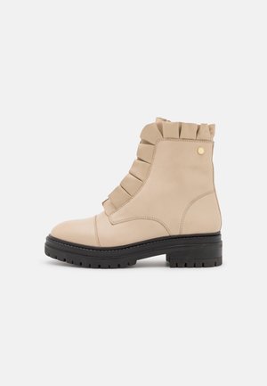 Beige leren enkellaarsje met gerimpelde details, zwarte rubberen platte zool, afgeronde neus en gouden accent aan de zijkant.