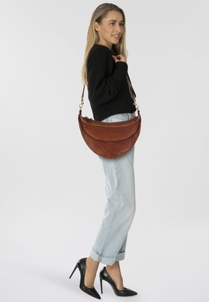 Bolso de piel marrón con forma redondeada, detalles de tachuelas y una correa de hombro desmontable. Modelo luciendo un atuendo casual con tacones altos.