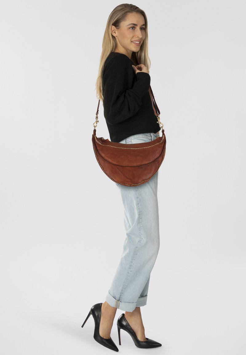 Braune Lederhandtasche mit runder Form, Nietenverzierungen und abnehmbarem Schulterriemen. Modell trägt ein lässiges Outfit mit hohen Absätzen.
