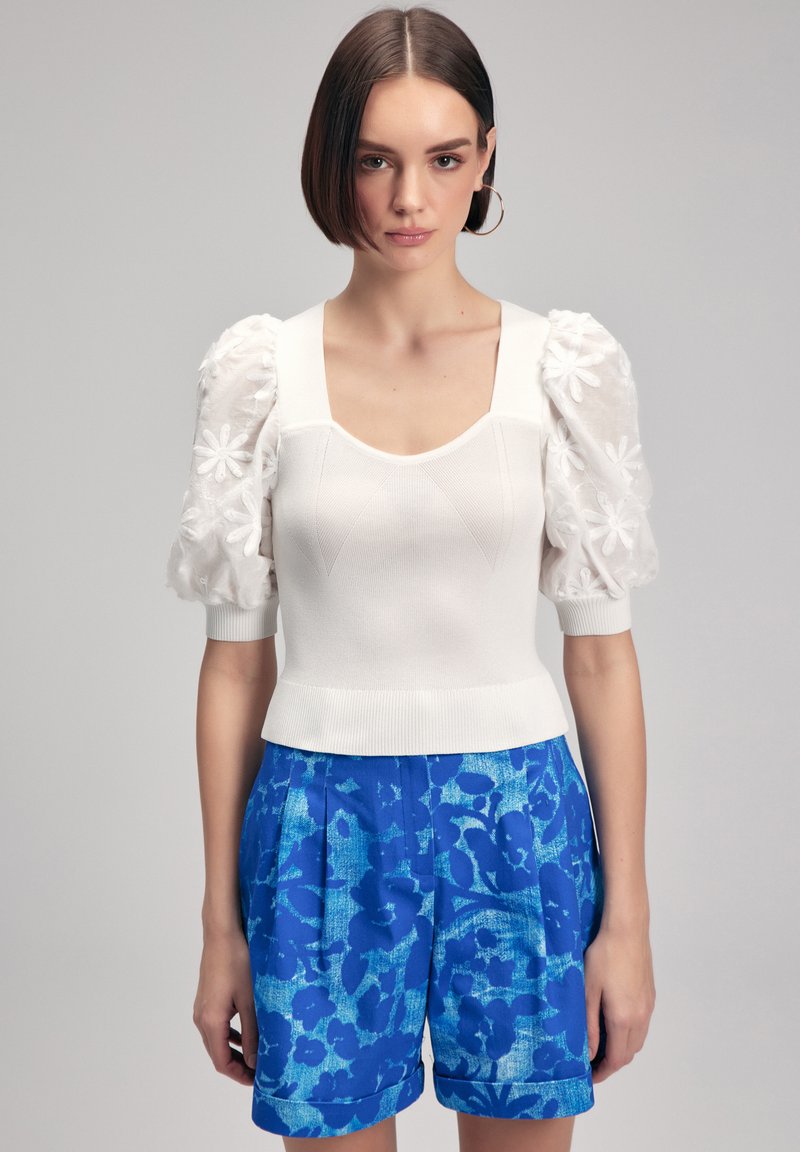 Top bianco a maglia con maniche a sbuffo con ricamo floreale, abbinato a pantaloni corti blu a fantasia. Il design include un collo quadrato e orlo a coste.