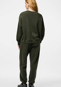 Moteris su ilgais blondiniais plaukais, apsirengusi tamsiai žalia per didelio dydžio džemperiu ir derančiomis sportinėmis kelnėmis, juodais sportbačiais, stovinti nugara į mus.