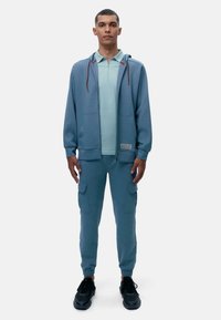 Svijetloplava kapuljača s crvenim vezicama, u kombinaciji sa odgovarajućim svijetloplavim polo majicom i cargo hlačama; ima džepove i rebraste manžete.