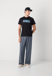 Camiseta de algodón negra con texto gráfico azul, combinada con pantalones deportivos grises de corte holgado. El modelo lleva una gorra negra y zapatillas ligeras.