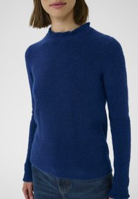 Modrý pletený sveter s textúrovaným povrchom, so zaobleným výstrihom a dlhými rukávmi, doplnený modrými džínsami.