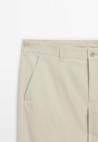 Pantalon beige montrant la ceinture, les passants de ceinture, la poche latérale et la fermeture à bouton sur un fond blanc uni.